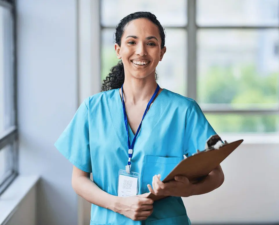 Smiling nurse holding clipboard in hospital hallway.